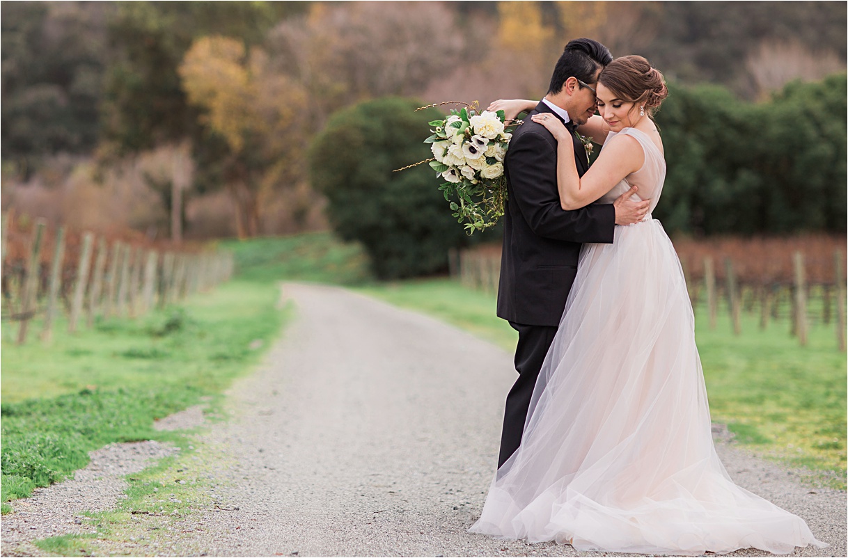 Bride and groom embracing on a dirt road lined by vineyards.